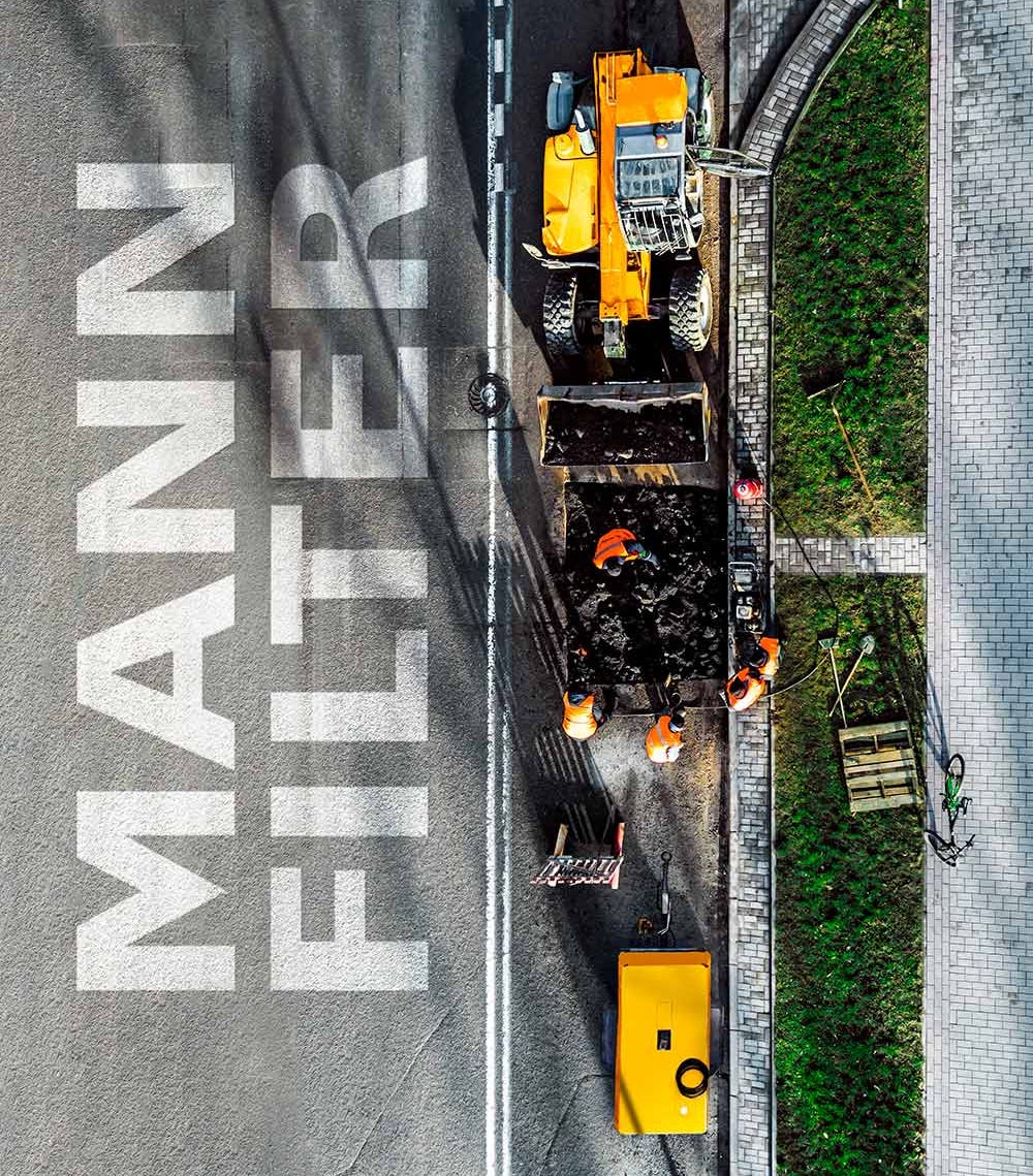 Blick auf eine Straßenbaustelle mit Fahrzeugen, Arbeitern und MANN-FILTER-Logo auf dem Boden