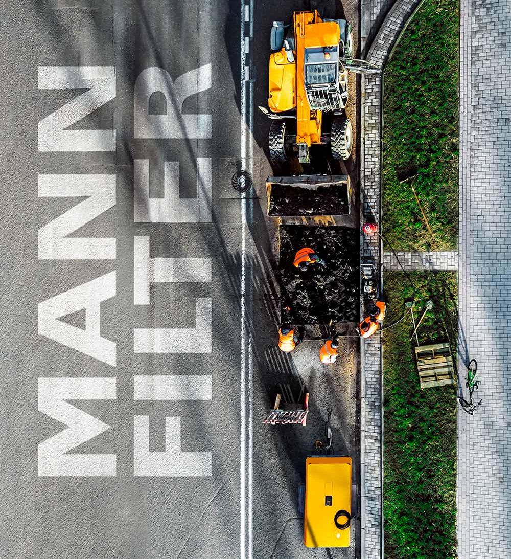 A view of a road construction site with vehicles, workers, and MANN-FILTER branding on the pavement
