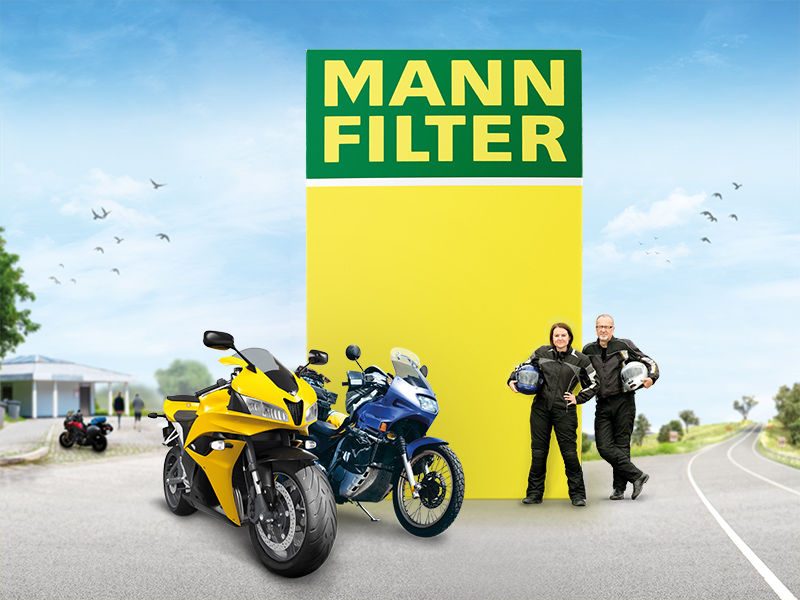 A group of motorcycles with riders near a bright yellow MANN-FILTER logo in an open spaceA group of motorcycles with riders near a bright yellow MANN-FILTER logo in an open space